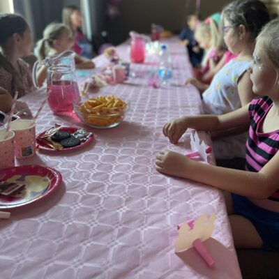 Na zdjęciu widzimy grupę dziewczynek, które siedzą przy stole i robią biżuterię.