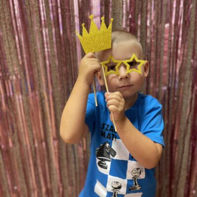 Zdjęcie przedstawia chłopca, który pozuje do zdjęcia. Do twarzy przykłada okulary gwiazdki i koronę "na patyku".