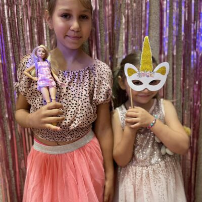 Zdjęcie przedstawia dwie dziewczynki pozujące do zdjęcia. Jedna z nich trzyma w ręku lalkę Barbie, a druga przykłada do twarzy maskę jednorożca "na patyku".