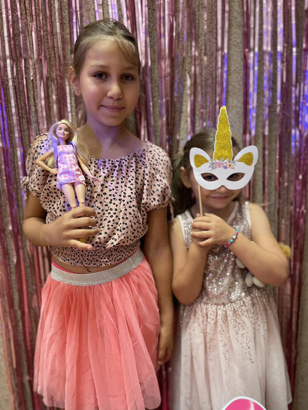 Zdjęcie przedstawia dwie dziewczynki pozujące do zdjęcia. Jedna z nich trzyma w ręku lalkę Barbie, a druga przykłada do twarzy maskę jednorożca "na patyku".