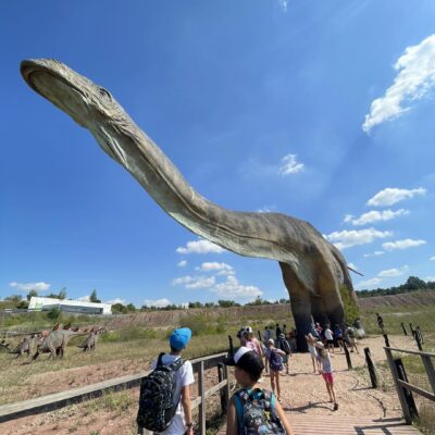 Zdjęcie przedstawia kilkoro dzieci podczas zwiedzania Juraparku. Dzieci podziwiają ogromnego dinozaura, który rozpościera swoją długą szyję ponad głowami zwiedzających.