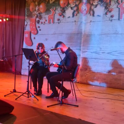 Młodzieżowy Zespół Wokalny na scenie podczas występu. Siedzą na krzesłach i grają na gitarze i skrzypcach.