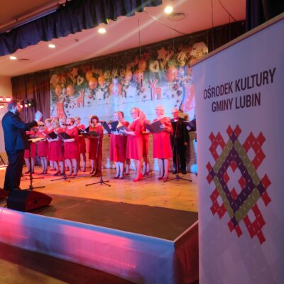 Chór Via Musica na scenie wraz z Dyrygentem. Kobiety w czerwonych sukniach dyrygent w czarnym fraku.