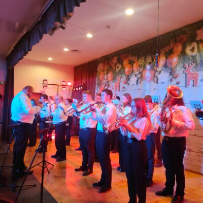 Młodzieżowa Orkiestra Dęta na scenie. Uczestnicy grają na trąbce, Flecie, Puzonie. Kapelmistrz na środku.