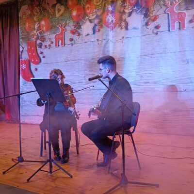 Młodzieżowy Zespół Wokalny na scenie podczas występu. Siedzą na krzesłach i grają na gitarze i skrzypcach.
