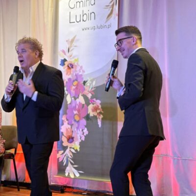 Dwóch mężczyzn w garniturach śpiewa na scenie do mikrofonów.