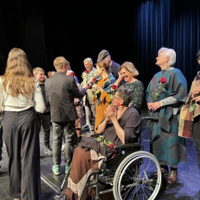 Na scenie teatru aktorzy na pierwszym planie kobieta na wózku. Zostają wręczone im kwiaty.