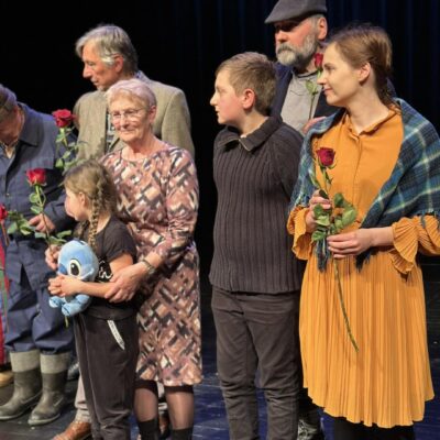 Aktorzy z wręczonymi kwiatami na pierwszym planie kobieta z różą i chłopiec w oddali inni aktorzy.