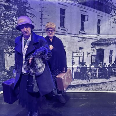 Aktorki teatru na scenie odgrywają rolę. Kobiety są ubrane i mają walizki na peronie.