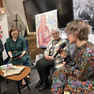 Trzy kobiety przy stole rozmawiają do mikrofonu. W tle obraz jednej z nich.