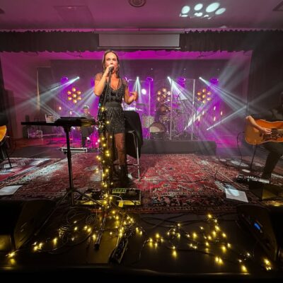 Kasia Kowalska na scenie. śpiewa do mikrofonu w czarnej sukience. Oświetlają ja światełka.