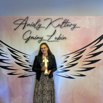 Edyta Szymańska z nagrodą na tle skrzydeł anioła pozuje do zdjęcia.