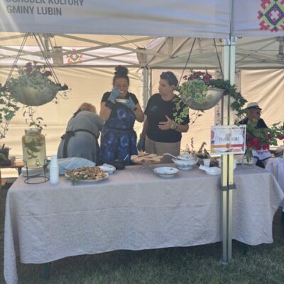 Dwie kobiety na stoisku, jedna próbuje zupę. Wiszą dekorację, kwiaty.