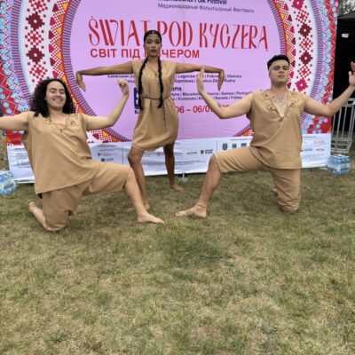 troje ludzi w strojach pozuje do zdjęcia na tle plakatu swiat pod kyczerą